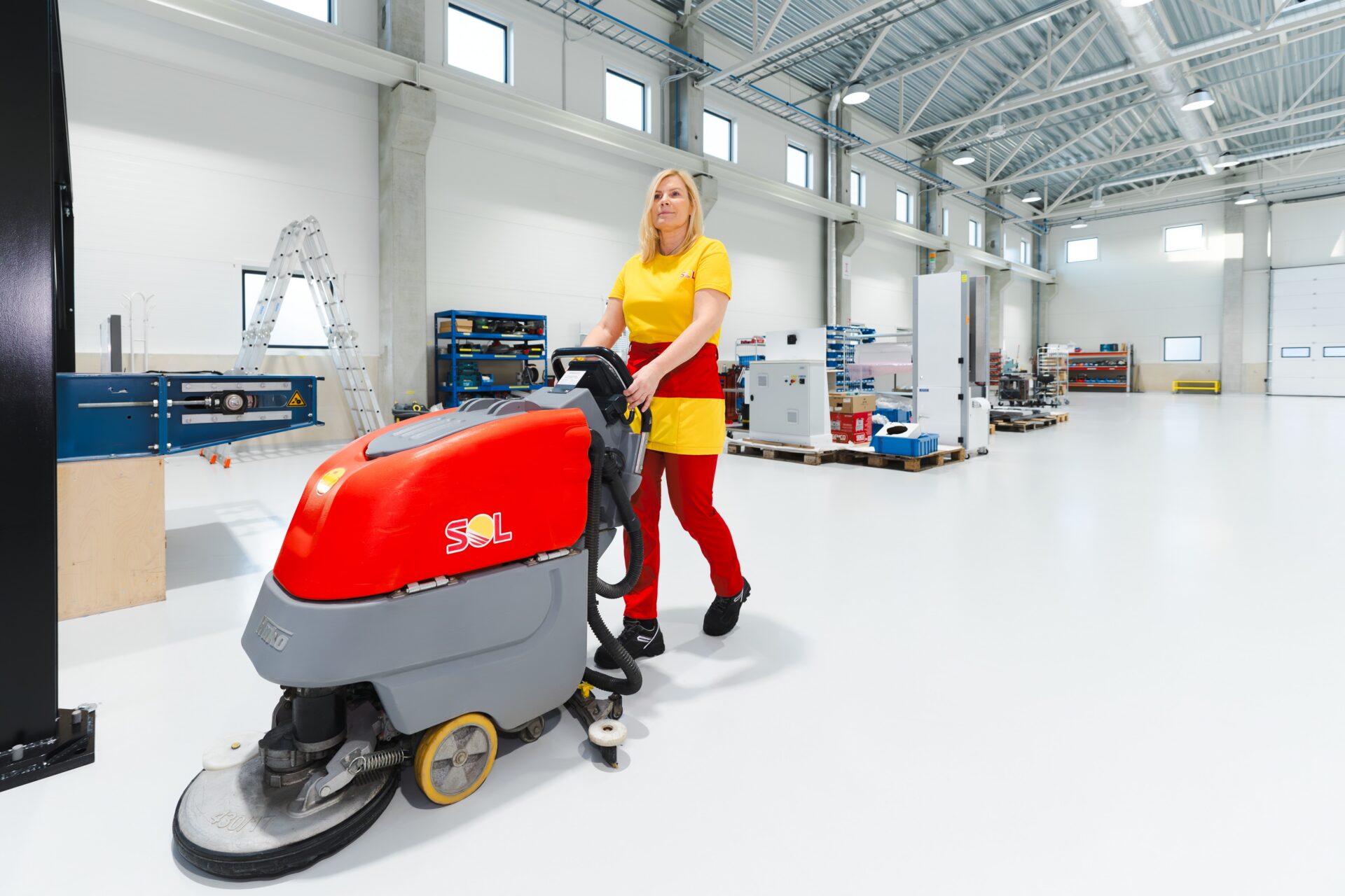 SOL darbiniece rūpīgi uzkopj industriālās telpas ar grīdas tīrīšanas iekārtu modernā ražošanas vidē.